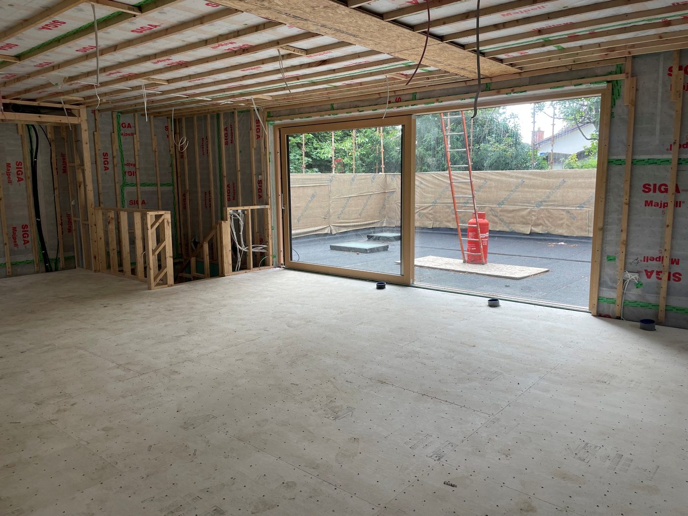 The interior of a house under construction, with exposed timber framing, taped airtightness membranes and a large glazed opening onto a rear terrace.